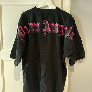 Svart Palm Angels t-shirt med rosa tryck - Äkta Svart oversized t-shirt från Palm Angels med stort rosa logotyp-tryck över ryggen och mindre logga i nacken. Klassisk rund hals och korta ärmar. Tillverkad i mjuk bomull, perfekt för en streetwear-look.