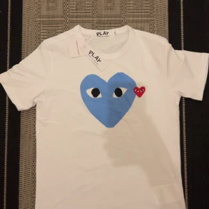 Vit Comme des Garçons Play t-shirt - Vit t-shirt från Comme des Garçons Play med stort blått hjärta med ögon och ett mindre rött hjärta på bröstet. Klassisk rund halsringning och korta ärmar. Ikonisk och stilren design som sticker ut.
