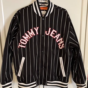 Jacka från Tommy Jeans - Snygg svart bomberjacka från Tommy Jeans med vita vertikala ränder och stora broderade bokstäver framtill. Jackan har ribbade muddar och krage i svartvitt, knappar framtill och två fickor. Perfekt för dig som vill sticka ut med en sportig och trendig look. Använd fåtal gånger och i väldigt fint skick!