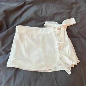 Vit omlottkjol från NA-KD x Pamela - Supersöt vit kort omlottkjol från NA-KD x Pamela. Kjolen har knytband i sidan och volangdetalj längs kanten för en extra feminin touch. Perfekt för dig som gillar en clean och fräsch look. Lätt och flowy känsla, passar till många olika stilar. Den är uppsydd och passar någon i xxs/xs