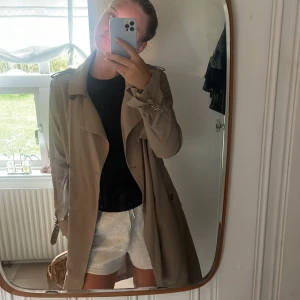 Beige trenchcoat med bälte och knappar - Snygg beige trenchcoat med klassisk krage, dubbelknäppning och bälte i midjan. Jackan har långa ärmar med spänne vid handlederna och axelklaffar. Perfekt för lager-på-lager och stilren look.