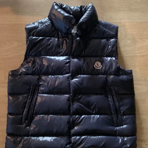 Blå dunväst från Moncler - Snygg blå dunväst från Moncler med hög krage och två dragkedjefickor på sidorna. Västen har ett glansigt yttertyg och klassisk Moncler-logga på bröstet. Perfekt för lager-på-lager och stilren look.