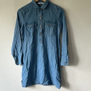 Jeansklänning från Mango - Säljer en klassisk jeansklänning från Mango i ljusblå denim. Klänningen har lång ärm, två bröstfickor med knapp, krage och knäppning framtill. Perfekt för en avslappnad och cool stil.