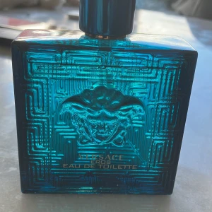 Versace Eros Eau de Toilette herrparfym - Versace Eros Eau de Toilette i en stilren, fyrkantig turkosblå glasflaska med Medusa-huvudet i relief på framsidan. Flaskan har guldiga detaljer på korken och ett geometriskt mönster runt om.