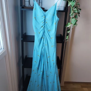 Blå Adoore klänning storlek 36 - Härlig ljusblå Adoore klänning med smala, reglerbara axelband och ett blommigt mönster i orange och grönt. Klänningen har en v-ringning och rynk framtill. Storlek 36. Mycket fint skick.