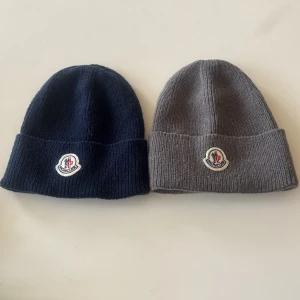 Moncler mössa i blå och grå - Två klassiska Moncler mössor i ribbstickad ull, en i marinblått och en i grått. Båda har Moncler-loggan framtill. Perfekt för kyliga dagar och enkel att matcha med olika outfits.  1 för 100kr