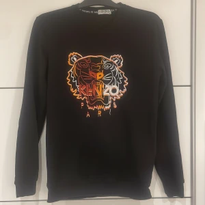 Svart sweatshirt med tigerbrodyr från Kenzo - Säljer en svart sweatshirt från Kenzo med ikonisk tigerbrodyr i orange, vitt och gult på bröstet. Tröjan har rund halsringning och långa ärmar. Perfekt för dig som gillar streetwear och vill sticka ut med en snygg detalj.