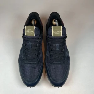 Valentino Garavani sneakers i mörkblå mocka och mesh - Säljer ett par snygga Valentino Garavani sneakers i mörkblå mocka och mesh med svarta snören och beige detaljer vid hälen. Skorna har en platt sula och rund tå, perfekt för dig som gillar stilrena och exklusiva sneakers.