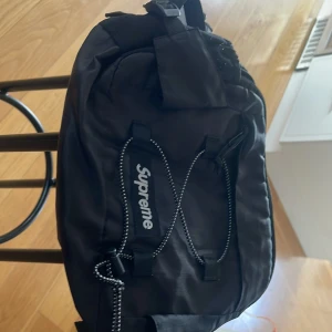 Svart axelväska från Supreme - Säljer en svart axelväska från Supreme med vit logga på framsidan. Väskan har justerbar rem, dragkedja och flera fack. Perfekt för att bära med sig det viktigaste och har en sportig streetwear-look.