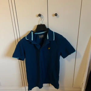 Blå pikétröja från Lyle & Scott - Snygg blå pikétröja från Lyle & Scott med ljusblå detaljer på kragen. Klassisk passform och korta ärmar, perfekt för en stilren look. Ikonisk gul logga på bröstet och mjukt bomullsmaterial.
