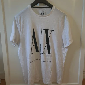 Vit t-shirt från Armani Exchange - Säljer en vit t-shirt från Armani Exchange med stor svart AX-logga framtill. Klassisk rund halsringning och normal passform. Perfekt för dig som gillar stilrena och enkla plagg med tydlig logga.