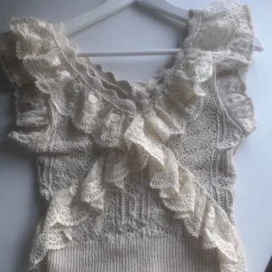 Beige stickad topp med spetsvolang från Zara - Supersöt beige stickad topp från Zara med breda spetsvolanger längs ringningen och axlarna. Perfekt till sommaren! Endast använd ett fåtal gånger.