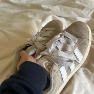 Beige Adidas Campus sneakers - Säljer ett par beige Adidas Campus sneakers med klassiska vita ränder på sidorna och vit sula. Skorna har snörning och är i mocka. Perfekta till i sommar! Observera att de har lite smuts längst bak vid insidan precis underfotleden, men det är inget som stör helhetsintrycket💕