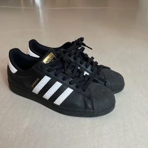 Adidas Superstar svartvita sneakers - Klassiska Adidas Superstar sneakers i svart med vita ränder och detaljer. Skorna har snörning, rund tå och platt sula. Perfekta för dig som gillar en tidlös streetstil och vill ha ett par ikoniska sneakers i garderoben. Bara använda några gånger men säljer för att de inte passar längre. Skorna är i väldigt bra skick👍🏻.