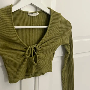 Olivgrön ribbad croppad topp från Pull&Bear - Trendig olivgrön croppad topp från Pull&Bear med långa ärmar och v-ringning. Toppen har ribbat material och snygg knytning framtill som ger en cool detalj. Perfekt att styla med jeans eller kjol för en avslappnad look.