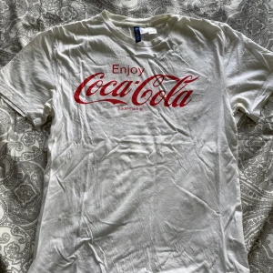 Vit Coca-Cola t-shirt från H&M Divided - Vit t-shirt från H&M Divided med röd Coca-Cola-logga och texten 'Enjoy Coca-Cola' på bröstet. Klassisk passform och rund halsringning. Perfekt för dig som gillar retrostil och ikoniska tryck.