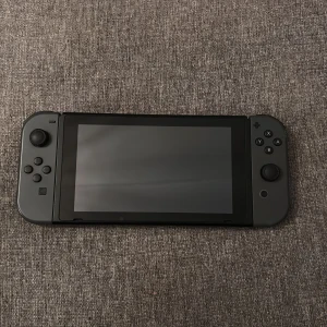 Nintendo Switch med fodral och spelkortsförvaring - Säljer en Nintendo Switch med grå Joy-Con och tillhörande svart hårt fodral med röd Nintendo Switch-logga. Fodralet har plats för konsolen och extra spelkort. Perfekt för gaming hemma eller på resan. Kommer med Fifa 21 och Pokemon Let’s go Pikachu