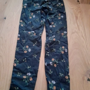 Mörkblå blommiga byxor - Säljer ett par mörkblå byxor med blommigt mönster i orange, lila och grönt. Byxorna har raka ben, hällor för bälte och fickor bak. Materialet är polyester och bomull, vilket gör dem mjuka och bekväma. Sparsamt använda på grund av för små. De har ytludd som detta material snabbt får. De är lite glansiga. De påminner lite om satin. Men mer matta