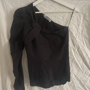 Svart oneshoulder-top från Zara - Säljer en svart oneshoulder-top från Zara med lång ärm på ena sidan och en snygg knutdetalj vid axeln. Toppen är tillverkad i en linneblandning och har en stilren, elegant look.