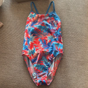 Färgglad baddräkt från Funkita - Säljer en baddräkt från Funkita med tropiskt blommigt mönster i rött, blått och inslag av vitt. Modellen har smala axelband och öppen rygg ( singel strap). Perfekt för simträning eller strandhäng. Använd fåtal gånger. Strl 14 år, 164.