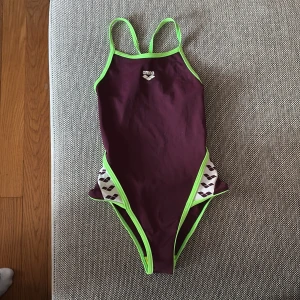 Baddräkt från Arena med gröna detaljer - Säljer en sportig baddräkt från Arena i mörklila med neongröna kanter och axelband. Sidorna har vita partier med svarta mönster. Perfekt för simträning eller bad. Snygg och bekväm passform med öppen rygg. Näst intill oanvänd. Ser ny ut. Storlek 10-11 år ca strl 140-148.