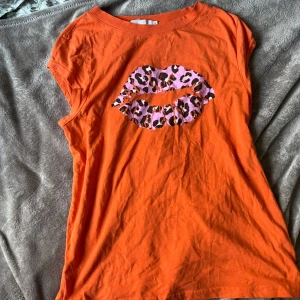 Orange t-shirt med leopardmönstrade läppar - Säljer en orange t-shirt med ett stort tryck av läppar i leopardmönster på bröstet. T-shirten har korta och öppna ärmar och rund halsringning. Perfekt för dig som vill sticka ut med färg och mönster! Ganska y2k!