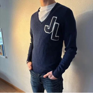 Mörkblå v-ringad J.Lindeberg - Säljer en mörkblå v-ringad tröja med stor vit JL-logga på bröstet. Tröjan har långa ärmar och ribbade muddar vid ärmslut och nederkant. Perfekt för en stilren och avslappnad look.