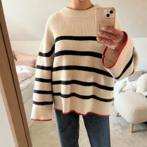 Randig oversized stickad tröja med röda detaljer - Säljer en krämvit stickad tröja med svarta horisontella ränder och röda detaljer vid ärmslut och nederkant. Tröjan har en oversized passform, rund hals och långa, vida ärmar. Perfekt för en avslappnad och trendig look.