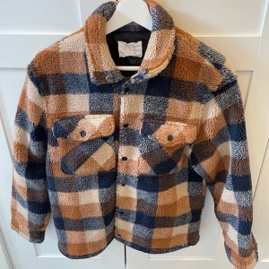 Rutig teddyjacka från Zara - Supermjuk och mysig teddyjacka från Zara i storlek S. Jackan har rutigt mönster i beige, blått och brunt, två bröstfickor med knappar och klassisk krage. Perfekt för kyliga dagar när du vill vara både varm och snygg.