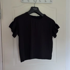 Svart ribbad topp med volangärmar - Säljer en svart ribbad topp med korta volangärmar. Toppen har rund halsringning och är tillverkad i ett stretchigt material som ger en snygg passform. Perfekt att styla till jeans eller kjol.