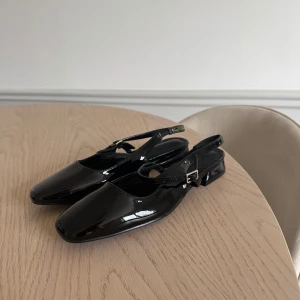 Svarta lackade ballerinaskor med rem - Säljer ett par svarta ballerinaskor i lack med smal rem och liten klack. Skorna har en klassisk stil med spänne vid ankeln. Perfekta för både vardag och finare event. Skorna är aldrig använda och i storlek 38!