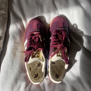 Adidas Samba OG Maroon  - Säljer ett par Adidas Samba OG sneakers i vinrött skinn med vita detaljer och klassiska tre ränder på sidan. Skorna har gummisula, snörning och guldfärgad Samba-text på sidan. De är helt nya och oanvända