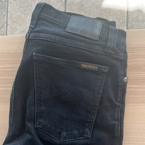 Svarta jeans från Nudie Jeans - Säljer ett par svarta jeans från Nudie Jeans med klassisk femficksdesign och subtila sömmar på bakfickorna. Jeansen har en rak passform och är tillverkade i ett mjukt denimtyg. Perfekta för dig som gillar stilrena och tidlösa jeans.
