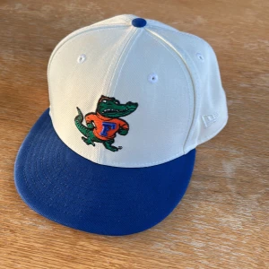 Vit och blå keps från New Era College Vault - Snygg keps från New Era College Vault med vit krona och blå skärm. Framtill finns ett broderat alligator-märke och texten Gators på sidan. Kepsen är i modellen 59FIFTY och är tillverkad i polyester. Sällsynt endast tillgänglig i USA 57,7 CM College football amerikansk
