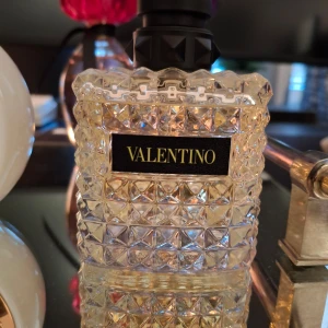 Valentino parfymflaska Yellow Dream - Säljer en lyxig parfym från Valentino i en transparent glasflaska med nitar och svart etikett. Flaskan har en fyrkantig form och ett svart lock med dekorativa detaljer. Perfekt för dig som gillar exklusiva dofter och snygg design.
