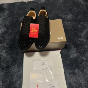 Svarta sneakers med nitar från Christian Louboutin - Säljer ett par svarta sneakers från Christian Louboutin med mattsvarta nitar på tån och klassisk röd sula. Skorna har snörning och är tillverkade i skinn med en stilren och lyxig design. Perfekta för dig som vill sticka ut med exklusiva detaljer.