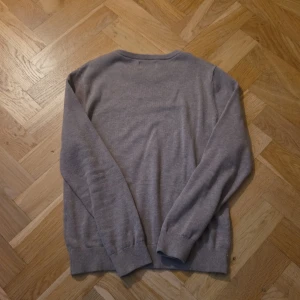 Beige stickad tröja från Lexington - Säljer en klassisk beige stickad tröja från Lexington med rund halsringning och diskret logga på bröstet. Tröjan har långa ärmar och ribbade muddar vid ärmslut och nederkant. Perfekt för en stilren och avslappnad look.