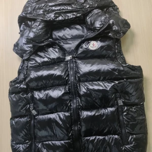 Svart dunväst från Moncler med huva - Snygg svart dunväst från Moncler med glansig finish och avtagbar huva. Västen har dragkedja framtill, två fickor med dragkedja och klassisk Moncler-logga på bröstet. Perfekt för lager-på-lager och stilren look. Man kan även scana moncler loggan