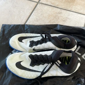 Nike Mamba spikskor  - Säljer ett par Nike Mamba löparskor i vitt med svarta detaljer och blå sula. Skorna har snörning och är tillverkade i ett lätt syntetmaterial med ventilerande mesh. Perfekta för dig som vill ha snabba och lätta skor till löpning.