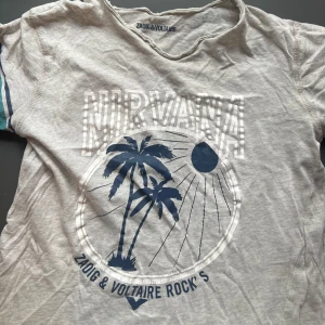 Grå t-shirt med tryck från Zadig & Voltaire - Snygg grå t-shirt från Zadig & Voltaire med tryck av palmer, sol och texten 'Nirvana' på framsidan. T-shirten har korta ärmar med blå och turkosa ränder samt broderad logga på ärmen.