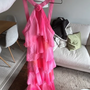Rosa volangklänning från Ellos - Säljer en superfin rosa långklänning från Ellos med flera lager volanger och en dekorativ blomdetalj vid axelbanden. Klänningen har ärmlös design och en härlig flowy känsla, perfekt för speciella tillfällen.