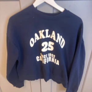 Mörkblå sweatshirt med Oakland-tryck - Mörkblå sweatshirt med vitt tryck framtill där det står 'Oakland 25 California'. Tröjan har rund halsringning och långa ärmar. Perfekt för en avslappnad stil.