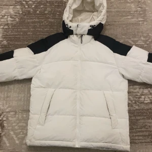 Vit och svart pufferjacka med avtagbar huva - Säljer en vit pufferjacka från jack&jones Man med svarta detaljer på axlar och ärmar. Jackan har avtagbar huva, dragkedja framtill och två stora fickor. Perfekt för kyliga dagar och riktigt skön att bära.