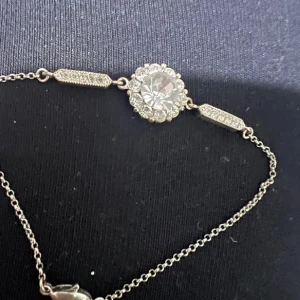 Silverfärgat armband med glittrande stenar - Ett elegant armband i silverfärg med en stor rund glittrande sten i mitten, omgiven av mindre stenar. Kedjan är tunn och smycket har dekorativa detaljer med små stenar på sidorna. Perfekt för att ge en extra touch till din outfit.