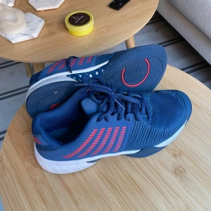 K-Swiss padelskor  - Säljer ett par snygga blå sneakers med röda ränder på sidorna och vita sulor. Skorna har snörning och en sportig design som passar perfekt till vardags eller träning. Bekväma och lätta att bära.