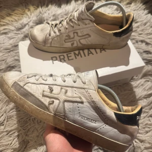 Premiata skor - Säljer ett par vita sneakers från Premiata med svarta detaljer på hälen och grå inslag vid sulan. Skorna har snörning och är tillverkade i skinn med tydlig logga på sidan. Perfekta nu till varmare väder! Inga defekter förutom att den vänstra skon är limmar på insidan som man ser på bild 4, därav de billiga priset! Bara att skriva vid minsta fundering‼️