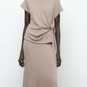 Beige omlottklänning med knyt i midjan - Stilren beige klänning med kort ärm och knyt i midjan som ger en snygg omlotteffekt. Klänningen har rak passform och är tillverkad i ett mjukt material. Perfekt för dig som gillar minimalistisk stil.