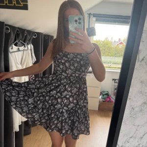 Svart blommig kortklänning - Säljer min favoritklänning då den inte passar längre! Det är en svart kortklänning med lila blommor. Klänningen har smala axelband, markerad midja och utställd kjol. Nypris: 399kr💜