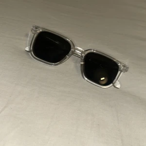 Transparenta solglasögon med svarta glas - Snygga solglasögon med transparent båge och svarta fyrkantiga glas. Ramen är i plast och har en modern, stilren look som passar till många outfits.