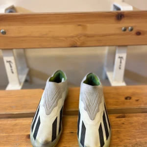 Adidas fotbollsskor med svart och grå design - Säljer ett par Adidas fotbollsskor i grått och svart med neongul sula. Skorna har en strömlinjeformad design utan snörning och är perfekta för dig som vill ha stilrena och lätta skor på planen.